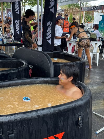 The image depicts an outdoor setting in บ้านสำนักตะแบก, Thailand. Several black inflatable tubs filled with a yellowish liquid and ice are lined up. A man is submerged in one of the tubs, his head and shoulders visible above the waterline. He has dark hair and appears to have some markings on his face. People are milling around in the background, some seated on white plastic chairs. One person is wearing leopard-print shorts and a matching top. A man in a white t-shirt and another man in an orange vest labeled "Official Camera" are also visible. Banners with a white logo on a black background are partially visible in the foreground and background. The environment appears to be a muddy area, likely a recovery zone or similar setup after an athletic event.