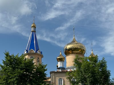 The media file captures an exterior view of a church in Cherven, Belarus, on a clear day. The main subjects are the church's towers and golden domes, set against a bright blue sky dotted with wispy white clouds. The architecture features light-colored brickwork, with a tall, slender tower topped with a blue, faceted roof and a golden spire. Adjacent to this, several rounded golden domes rise, some larger and more prominent than others, each crowned with a cross. Green trees with dense foliage partially obscure the lower sections of the church building and frame the scene. The sunlight suggests it is daytime, likely late morning or afternoon, given the brightness and the clear sky. There is a faint contrail from an airplane visible in the sky. No people or specific events are depicted. The image focuses on the serene and majestic appearance of the religious structure.