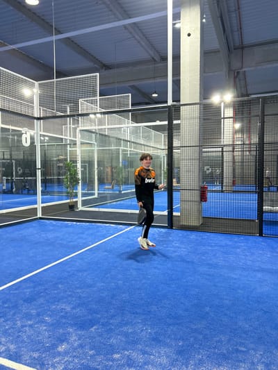A young person stands on a blue paddle court, holding a paddle. They are wearing black pants and a long-sleeved shirt with orange accents. The court is enclosed by clear glass and metal mesh fencing, with another court visible in the background. The lighting is bright, revealing a high ceiling and industrial-style architecture. The image is rotated slightly to the left.