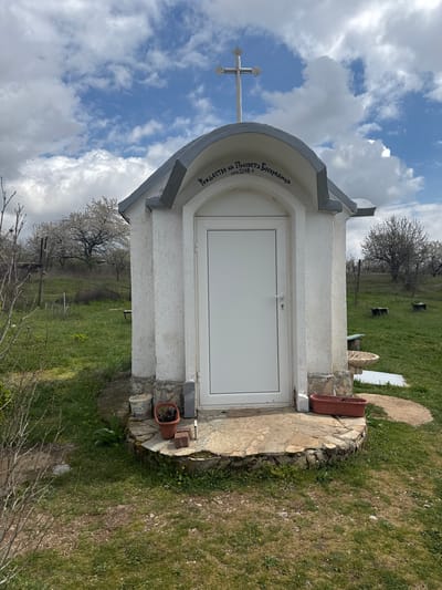 A small, white, door-covered structure with a cross on top stands on a grassy area with some trees in the background. The structure is identified by text above the door as the "Holy Mother of God" location in Gorna Grashtitsa, Bulgaria. Stone paths and planters surround the structure. The sky overhead is cloudy.