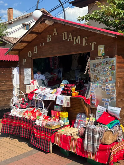 This is a full-shot, eye-level, outdoor photograph of a souvenir stall in Chavdar, Bulgaria. The stall is made of wood with a red roof and is open to display its wares. A sign above the stall reads "ПОД НАМЕТ" in raised letters. The stall is laden with various items including embroidered shirts, jars of honey, baskets of small gifts, textiles, and religious icons. There is a mannequin dressed in an embroidered shirt. A sign on the wall reads "ДЪРВЕНИ ИЗДЕЛИЯ" (wooden items). Another sign displays the number "7" and what appears to be a green graphic of a plant. On the right side of the stall, signs in Bulgarian are visible, including one that says "РАБОТНИ ТЕНИСКИ" (work shoes). The stall is set up on a brick-paved walkway. The background shows a building with a red roof and a chimney, and green trees against a bright blue sky with white clouds. The overall scene suggests a market or fair setting in a Bulgarian town.