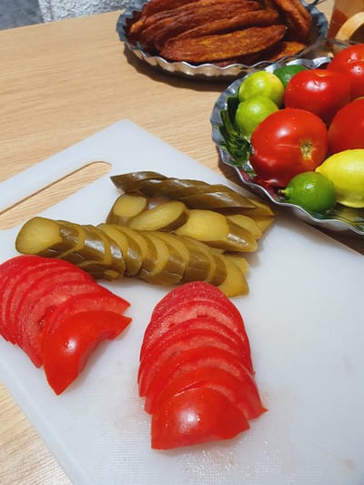 In Kheyrabad, Iran, a close-up, eye-level shot showcases a food preparation scene. The focus is on a white cutting board where sliced tomatoes and pickles are arranged. In the background, a silver bowl holds red tomatoes and green limes, while another metallic bowl contains fried food, possibly churros, with a textured surface. The subjects are purely edible items, and no people or interactions are visible. The environment appears to be a kitchen or dining area, with a wooden table surface visible. The lighting is natural, highlighting the textures and colors of the food.