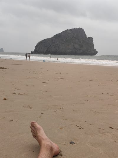 The scene depicts a beach in Hải Phòng, Vietnam, characterized by gray skies and a rocky island in the distance. The foreground is dominated by sand, upon which lies a human foot, likely belonging to the photographer. Further up the beach, there are at least 2 people walking along the shore. In the background, waves are visible, as well as a large dark, rocky island. The overall atmosphere of the scene appears to be overcast and tranquil.