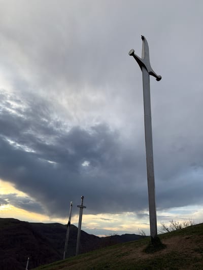 The image shows a landscape with a focus on several monumental metal swords erected vertically into the ground. The swords have a stylized appearance, with large, simplified blades and crossguards. The sky is overcast with dramatic, swirling clouds, ranging from light gray to dark gray hues. In the background, a hillside or mountain is visible, partially covered in vegetation. The location is the city of Zemo Nichbisi, Georgia.