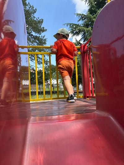 A young boy, wearing an orange t-shirt and orange shorts, stands on a playground slide, his back to the camera. He is wearing a green cap and black sneakers with yellow accents. The boy is holding onto a yellow railing. The slide is red, and the platform is made of wooden planks. In the background, there is a bright blue sky with some white clouds and green trees. The scene is taking place outdoors in the city of Vinarsko, Bulgaria.