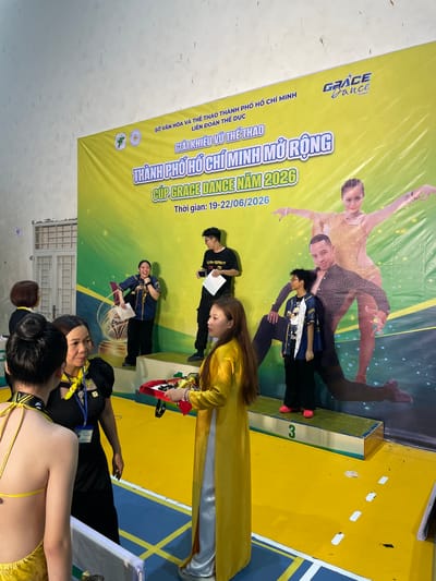 The image depicts an awards ceremony or competition taking place in Thuận An city, Vietnam. A large yellow banner serves as the backdrop, featuring text in Vietnamese and the logo "GRACE Dance Academy." The banner prominently displays the title "GIẢI KHIÊU VŨ THỂ THAO THÀNH PHỐ HỒ CHÍ MINH MỞ RỘNG - CÚP GRACE DANCE NĂM 2026" and the dates "19-22/06/2026."
In the foreground, several individuals are present. On the right, a person in a bright yellow áo dài is presenting a tray with trophies to a young woman. Another person, dressed in black with a yellow scarf, stands to the left of the presenter, holding a document. Behind them, a woman in a blue and gold jacket is laughing and holding papers. Further to the left, two young women are conversing, with one facing away from the camera and wearing a yellow garment. A young man wearing a black t-shirt and glasses stands on a podium labeled with the number "3" to the right of the banner. He holds a piece of paper. A large printed image of a dancing couple is visible on the banner behind the participants. The environment appears to be an indoor venue with a yellow floor marked with white lines.