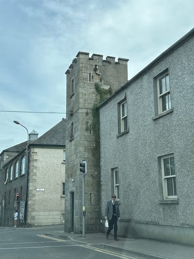 The image depicts a street scene in Carlow, Ireland, featuring a mix of architectural styles and a pedestrian. In the foreground, a man in a gray suit, dark tie, and a flat cap is walking on the sidewalk, carrying a white plastic bag. He is facing slightly towards the viewer. Behind him, a textured gray building with multiple windows lines the street. To the left of the building, a tall, stone tower with crenellations rises, possibly a historical structure, with ivy growing on its side. Further to the left, another building with arched windows is visible. A traffic light is positioned on the right side of the tower, currently showing red. In the background, the sky is overcast with scattered clouds. The street is paved with asphalt, marked with yellow lines. A street sign that reads "CHAFF ST" is affixed to one of the buildings. Another person, a woman in a light-colored dress, is standing near a traffic light and a signpost on the far left of the image.