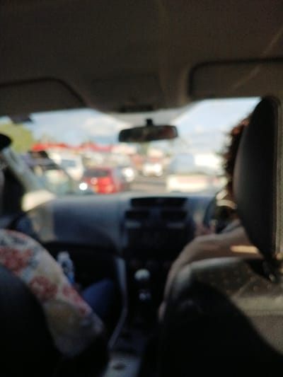 The image provides a first-person perspective from the rear passenger seat of a vehicle on an urban street in Harare, Zimbabwe. In the immediate foreground, the top of a dark headrest is visible on the right. To the left, the left shoulder and arm of a front passenger, clad in a light-colored, patterned garment, are partially visible. The vehicle's dashboard, steering wheel, and central console are present but appear out of focus. Through the windshield, a scene of dense daytime vehicular traffic is observed. Numerous blurred vehicles populate the street, prominently featuring a distinct red car among an array of other lighter-colored vehicles. The sky above the street is bright, indicating daylight conditions with indistinct cloud formations.