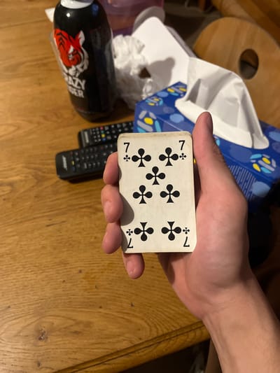 A light-skinned human hand holds a white playing card, the 7 of Clubs, vertically in the foreground, with the thumb gripping the left side. The card displays seven black club symbols and black '7' numerals in all four corners, showing minor wear along the edges. The scene takes place on a light brown wooden table surface. To the upper left, a black bottle with a red tiger logo and "CRAZY TIGER" text is visible. Two black remote controls are positioned behind the card, slightly to the left. Further right and behind the hand, a blue and white patterned tissue box contains a white tissue protruding from its opening, flanked by a crumpled white tissue to its left. The background is an indistinct indoor setting. The precise location within Amiens, France, cannot be determined from the visual information presented.