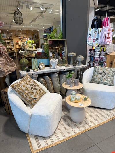 The image depicts a retail store interior, likely a home decor or gift shop, with a display setup in the foreground. Two plush white armchairs with textured upholstery are arranged on a striped woven rug. A patterned throw pillow, featuring a geometric design in brown and gold, is placed on the left armchair, while another similar pillow with green and beige tones sits on the right armchair. Between the chairs, a small, light-colored side table holds two green patterned mugs and a small glass bird figurine. Another, larger side table behind it displays three ceramic planters, one with a small plant, and two decorative vases.

Behind the seating area, a rustic wooden table is laden with various decorative items. Notable objects include a green desk lamp, a striped green vase, a golden oval picture frame, and a white sculptural piece resembling a cluster of spherical shapes. Further back, shelves are visible displaying a variety of items, including framed art, glassware, and potted plants. To the right of the display, a rack holds several tote bags with various whimsical prints, including cats and pretzels. In the background, the store environment is suggested by overhead lighting fixtures and glimpses of other merchandise and shelving. The scene is brightly lit, with a focus on the display in the foreground. There are no visible people in the immediate vicinity of the described display. The scene is staged for retail presentation.