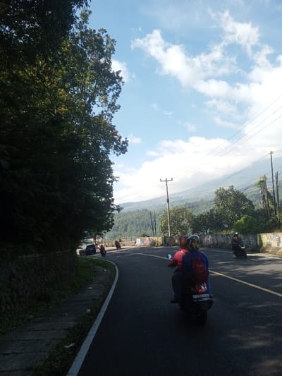 The image presents a daytime scene on an asphalt road curving right through a hilly, vegetated landscape, located in the city of Ka Thaung (Upper), Myanmar. The sky is clear blue with scattered white cumulus clouds. On the left, a dense mass of green foliage and trees covers a steep embankment, with a stone retaining wall visible at its base.
In the immediate foreground, the rear of a dark-colored scooter is visible, carrying two individuals away from the camera. The driver wears a red helmet and red shirt, while the passenger wears a blue helmet and a blue backpack. Further along the road curve, several other scooters are observed, each with one or two riders, also traveling in the same direction. A dark car is partially visible on the far left of the road ahead.
To the right of the road, a concrete barrier or railing, marked with graffiti, separates the road from a downward slope. Utility poles with power lines traverse the mid-distance. The background features extensive green mountains or hills, with distant settlements or cultivated areas discernible on the slopes. Sunlight illuminates the scene, casting shadows from the trees on the left side of the road.
