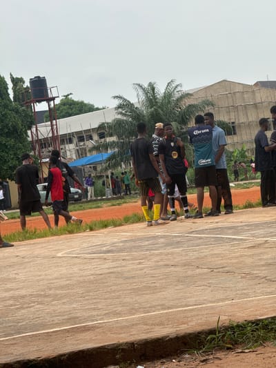 The image depicts a group of young men gathered on an outdoor basketball court in Amansee, Nigeria. The court surface is made of weathered wood with white court lines. To the right, a patch of green grass and reddish-brown dirt borders the court. In the background, palm trees and other lush greenery are visible, with a multi-story building undergoing construction or renovation. A large black water tank on a metal frame also stands out against the overcast sky.
The individuals are dressed casually, mostly in athletic wear. Some are wearing t-shirts with logos, including one that clearly reads "Corona" and another with "caliente.mx". Another shirt has a circular logo that appears to be a Spotify logo. The people are standing in small clusters, some facing each other in conversation, while others appear to be watching something or waiting. The overall scene suggests a casual gathering or a break during a sports activity. The weather appears to be overcast, contributing to a muted daylight. The presence of a basketball court, the attire of the individuals, and the youthful energy implied by the group suggest a sports-related context, possibly a pickup game or practice.
