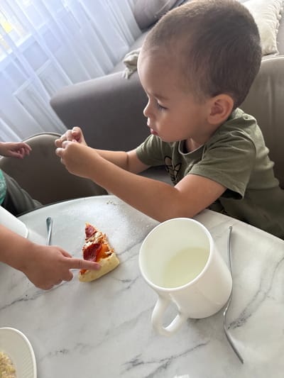 In the city of Nizhneyarkeyevo, Russia, a young boy wearing a green t-shirt is seated at a white marble table, looking down at a slice of pepperoni pizza. His hands are clasped in front of him. To his left, another child's hands are visible, with one child pointing at the pizza. A white mug filled with a light-colored liquid and a fork are also on the table. The background shows a blurred view of a couch and curtains.