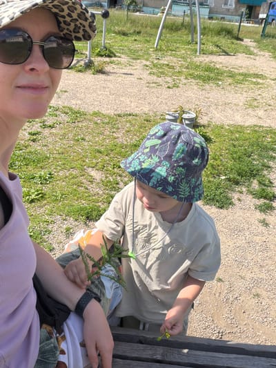 In Кульдур, Russia, a woman wearing a leopard-print sun hat and sunglasses is in the foreground, partially visible on the left. She is wearing a light purple sleeveless top. Beside her, a young child wearing a dark bucket hat with a green leafy pattern and a light beige t-shirt is seated. The child is holding a sprig of greenery and looking down. The scene is outdoors, with a gravelly path and patches of grass visible. In the background, there are some outdoor exercise equipment and buildings, suggesting a park or playground setting. The lighting indicates it is daytime.