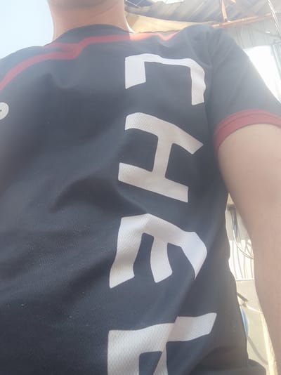 A close-up, chest-level shot focuses on a person wearing a dark-colored t-shirt with bold white lettering. The letters appear to spell out "CHE" vertically down the front of the shirt. There are red accents on the collar and sleeve edges. The fabric of the shirt shows texture from its mesh-like material. The background is blurred and appears to be outdoors with sunlight filtering from above, suggesting an open area or possibly a rooftop. The environment is consistent with an urban setting, specifically the city of Kobeliaky, Ukraine, given the lighting and potential background structures that could be part of a building or outdoor utility area. The image is taken from a slightly upward angle.