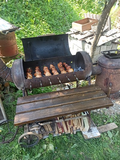 This is an outdoor scene in Kniazhdvir, Ukraine, featuring a makeshift barbecue grill constructed from a large metal barrel. The grill is open, revealing several skewers of meat cooking over hot coals. In the background, there is lush green foliage, a rusty barrel with a corrugated metal lid, and various construction materials, including bricks, roofing tiles, and metal boxes. A wooden bench is positioned in front of the grill. The overall environment appears to be a backyard or a rural setting. There are no people visible in the image.