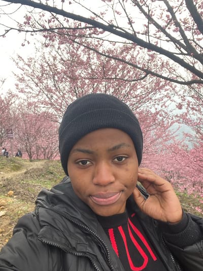 A young adult female of African descent is captured in a selfie taken outdoors in a park or scenic area within Caijia, China. She is centered in the frame, wearing a black knit beanie, a black puffer jacket, and a black sweater with a red patterned design. Her right hand is positioned at the side of her head, near her ear, and she displays a subtle smile. The background is dominated by a dense grove of pink-blossoming trees, consistent with cherry blossoms, extending across the midground and into the distance under an overcast sky. A dirt path is visible on the left side of the midground, where several indistinct individuals are seated among the trees. One group of people is situated near a partially obscured white rectangular sign displaying red characters. Hazy mountain silhouettes are discernible through the tree foliage in the distant background on the right.