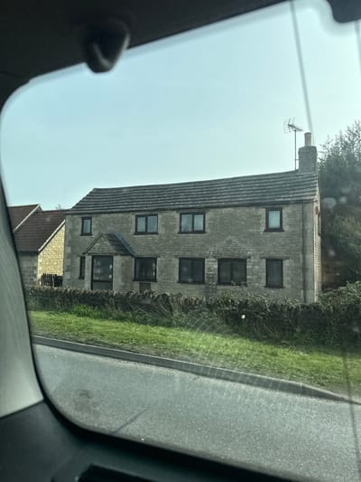 The image depicts a two-story, light-colored stone house situated in Thorpe Waterville, United Kingdom, viewed from inside a vehicle through a side window. The house features a dark gray tiled roof, a prominent stone chimney stack on the right side from which a multi-element television antenna extends upwards, and nine dark-framed windows (eight on the main facade, one on the right side). A small, stone-built porch with a darker roof covers the central entrance on the ground floor. To the left of the main house, part of another stone building with a reddish-brown tiled roof is visible. In the foreground, an asphalt road with a curb transitions into a grassy verge. A low dry-stone wall, partially obscured by overgrown green vegetation, separates the verge from the house's property. A single white flower is visible within the vegetation in front of the wall. The sky above is clear and light blue. The dark interior trim of the vehicle's window frames the scene, with a dark object partially visible at the top center. No individuals or active interactions are depicted.