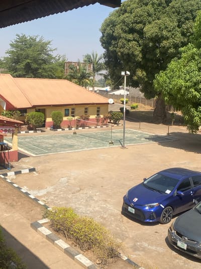 This image depicts an outdoor compound, likely a school or recreational facility, located in Tudun Nupawa, Nigeria, under bright daylight conditions. The scene is viewed from an elevated position, with a roof overhang visible at the top left.
In the mid-ground, a single-story building with yellowish walls and a red-brown corrugated metal roof extends horizontally. It features multiple rectangular windows and is fronted by landscaped shrubs. To its left, a partial view of another structure with a similar roof color displays a sign partially legible as "N-CHICKENS."
Immediately in front of the main building, a rectangular court, painted light green with white boundary lines, is visible. A net is strung across its center, indicative of a badminton or volleyball court. A tall, white metal pole equipped with floodlights stands adjacent to the court on the right. Further right, a satellite dish is mounted on a pole near the building.
The ground surrounding the court and structures is primarily unpaved dirt or a worn concrete surface. Black and white painted curbs define edges of pathways and a small, shrub-filled planter in the foreground.
Two sedans are parked in the lower right quadrant. The foremost vehicle is a dark blue Toyota Corolla, displaying an "L" plate (learner's permit) on its front bumper and windshield. Its license plate originates from "KANO." Partially obscured behind it is a darker-colored sedan, also with a "KANO" license plate.
The background features numerous green trees, including a very large specimen on the right. A raised, rectangular structure, possibly a water tank, is visible among the trees further back. A block wall delineates the property perimeter in the distance. No individuals are visible in the scene.