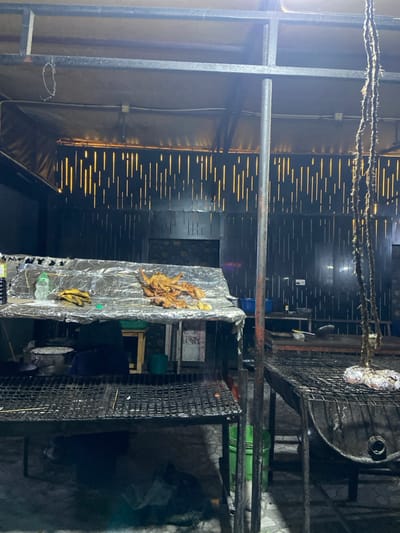 The image shows a dimly lit outdoor cooking area, likely a street food stall, located in Dodoma, Tanzania, at the coordinates -6.157 latitude and 35.723 longitude. There are two large metal grills, one on top of the other, both with a layer of charcoal embers and ash. One grill is topped with a sheet of aluminum foil, holding cooked chicken and possibly bananas. A third, larger, cylindrical grill sits on the right, also with charcoal embers, and a piece of meat is visible on the grill itself. The scene is messy, with evidence of cooking and food preparation. There are also several unidentified black objects in the scene, potentially containers for cooking ingredients or tools. The walls of the cooking area are black with a patterned design and covered by a canopy. The floor is a rough, uneven surface. There are no people present in the image.