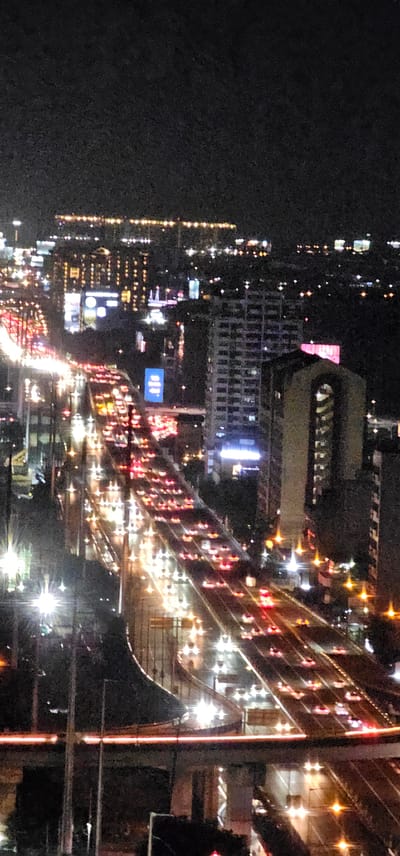 The image shows a nighttime aerial view of a busy highway in the city of Makati, Philippines. The highway is filled with cars, their headlights and taillights creating streaks of white and red light that illuminate the scene. Tall buildings, lit with numerous windows, flank the highway, indicating a densely populated urban environment. Some of these buildings appear to be commercial or residential high-rises. The overall impression is one of a bustling city at night, characterized by heavy traffic and illuminated architecture.