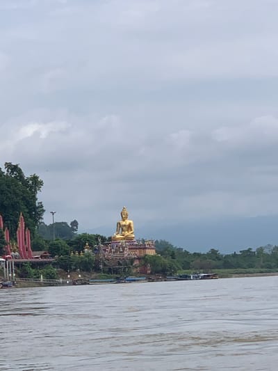 The image captures a wide view of the Mekong River in Chiang Saen District, Thailand, on an overcast day. On the riverbank, a prominent golden Buddha statue sits atop a structure, overlooking the water. The statue is seated in a meditative pose. Behind the Buddha, lush green trees and some traditional Thai architecture, including red and gold spires, are visible. Several boats are docked along the riverbank, with some appearing to be longtail boats. The river itself is a muddy brown, with visible ripples suggesting some movement. The sky is filled with grey clouds, hinting at potential rain. The overall scene suggests a tranquil yet culturally rich riverside setting.