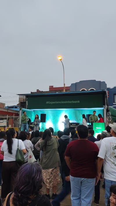 The image depicts a live event, likely a performance, taking place on what appears to be a mobile stage or vehicle in an urban setting. A crowd is gathered to watch. The vehicle, identified with the logo or slogan "#ExperienciasInterbank", suggests a promotional event by the Interbank financial institution. The event seems to be taking place outdoors, and the lighting indicates it could be dusk. A street lamp is illuminated nearby. Buildings are visible in the background. Based on other data I have, the location is Barranco, Peru.