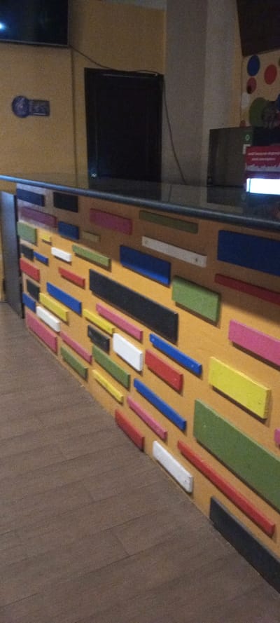 An indoor scene in Cipanas, Indonesia, displays a reception or service counter viewed from a low, oblique angle. The foreground features a dark brown wood plank floor. A counter, approximately waist-height, dominates the midground. Its top surface is a reflective black. The front of the counter is decorated with numerous rectangular wooden blocks of various sizes and orientations, painted in colors including white, red, blue, green, yellow, pink, and black, affixed to a light orange or tan panel. A dark vertical panel flanks the left side of the counter. In the background, a light orange wall is visible. A flat-screen television is mounted high on the left wall section. Below it, a dark, rectangular object is affixed to the wall. To the right, a light-colored structural pillar ascends. Further right, a wall section is adorned with circular shapes in red, blue, and green. On the black counter surface, a dark monitor or screen is positioned, and to its right, a partially visible illuminated screen displays red and white graphical elements. No individuals or active interactions are present.