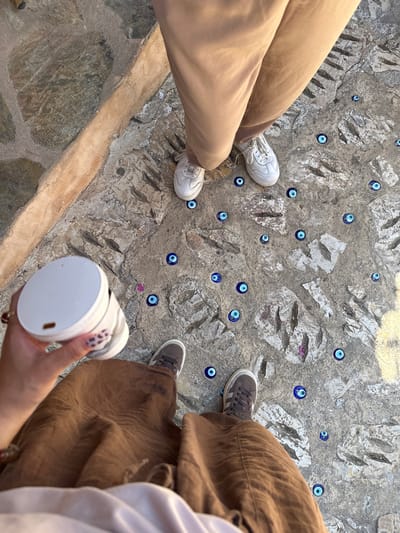 The image, captured from an overhead perspective, shows the lower bodies of two individuals standing on a textured stone pathway in Kaş, Turkey. The ground is composed of rough, irregularly shaped grey and off-white stones, with numerous small, round blue and white evil eye charms (nazar boncuğu) embedded or scattered across the surface, a common cultural motif in the region.

On the left, one person wears loose-fitting beige trousers and white athletic sneakers with distinct lacing. Their legs are positioned slightly apart, facing towards the lower left. On the right, the second person wears wide-legged, flowing brown trousers and grey sneakers with white soles and trim. This individual's right hand is visible in the lower right corner, holding a white cylindrical object, possibly a camera lens, portable speaker, or a thermos. A ring is visible on a finger, and a reddish bracelet is on the wrist.

The lighting indicates daytime conditions, with natural light casting subtle shadows, suggesting a clear sky. The rough, earthy texture of the stone path and the presence of the evil eye charms provide strong location cues, aligning with the provided context of Kaş, Turkey, often found in traditional or historical areas. There is no visible text in the image. The scene conveys a casual moment, perhaps during sightseeing or a walk.