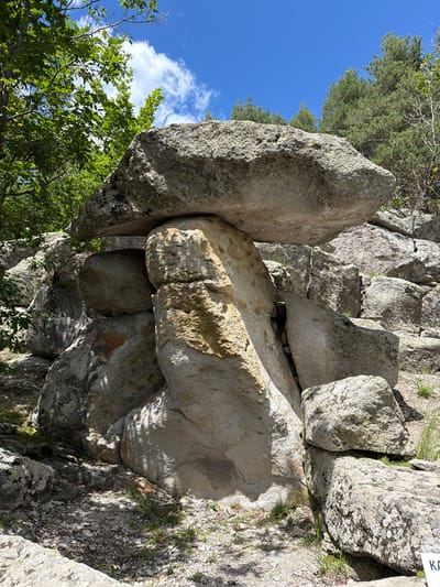 The image shows a large, ancient dolmen-like rock formation in the Laki region of Bulgaria. The formation consists of several massive, irregularly shaped stones. A large, flat stone rests precariously atop a cluster of upright stones, creating a cavernous opening beneath. The surrounding environment is a natural, rocky landscape with trees and vegetation. The sky is blue with scattered clouds. The lighting suggests it is daytime, possibly late morning or early afternoon, with sunlight casting shadows across the rocks and ground. The overall impression is one of natural geological formation or ancient human construction in a wild setting.