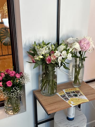 The image shows a close-up of a display featuring floral arrangements and promotional flyers. In the foreground, on the left, a glass vase holds a bouquet of pink roses interspersed with white baby's breath. To the right of this, a larger glass vase contains a mixed arrangement of white lilies and pink roses. Behind these, to the right, a second, taller glass vase holds white and pink flowers. A wooden shelf, supported by a metal frame, holds the two right-hand vases and two yellow flyers. The flyers have the word "ARTS" prominently displayed and appear to be advertisements for an arts studio. The background is a plain, light-colored wall. A portion of a mirrored surface with a dark frame is visible on the far left, reflecting a Monstera plant. The overall impression is that of a welcoming display, possibly in a studio or event space, in Zakharovo, Russia.