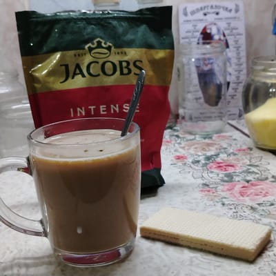 A clear glass mug filled with a light brown, frothy beverage sits on a tablecloth patterned with pink and white flowers. A dark gray spoon is partially submerged in the drink. To the right of the mug is a wafer cookie with visible layers. In the background, a bag of Jacobs Intense coffee is visible, featuring a gold and dark green design with the brand name "JACOBS" in large black letters and the word "INTENSE" below. Behind the coffee bag, there are glass jars and a small, white printed paper with some text. The overall setting appears to be a domestic kitchen environment. There are no individuals present in the image. The scene does not provide any information about the location in Ekibastuz, Kazakhstan.
