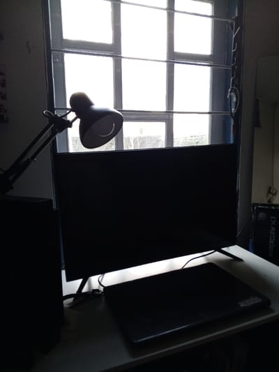 The image displays an interior room setup, specified to be located in Villanueva de Gállego, Spain. A large, multi-pane window with a light-colored frame dominates the upper half of the scene, admitting strong, blown-out white light from the exterior and creating silhouettes of the foreground objects.
On a light-colored desk occupying the lower portion of the frame, several electronic items are arranged. To the left, a black, articulated desk lamp with a conical shade is positioned, angled towards the right. Centrally, a large, dark, flat-screen monitor or television stands on the desk, its screen black, indicating it is off or unpowered. A closed, dark-colored laptop rests on the desk in front of the monitor, slightly to the right. To the far left of the desk, a dark, vertical rectangular object, likely a computer tower or speaker, is visible. On the far right, a partial view of a dark rectangular object bearing white text "HYPERCLAW X1" is present. Multiple dark cables are visible on the desk surface, connecting the devices.
The wall to the immediate right of the window appears darker, possibly a door frame, from which a string of dark beads and a metallic carabiner-like object hang vertically. No individuals are present in the scene.