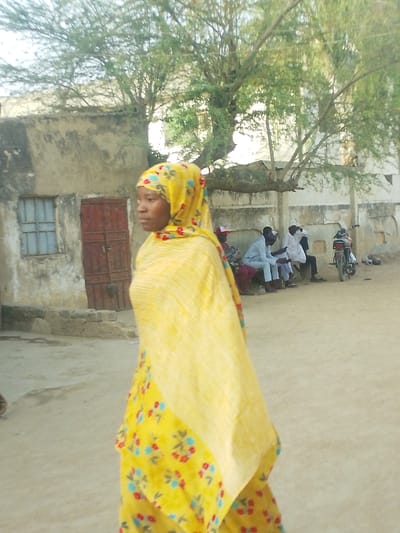 A daylight scene in Garun Gawa, Nigeria, depicts a woman in the foreground walking left, her left profile visible. She wears a yellow hijab and a matching dress with red and blue floral patterns. In the mid-ground left, a light-colored building with a weathered facade features a small barred window and a closed dark red wooden door. To the mid-ground right, approximately five men are seated on a low structure against a lighter wall, appearing to interact. One man wears a red and white cap. A motorcycle is parked on the far right. The environment consists of unpaved dirt ground and deciduous trees with sparse foliage overhead.