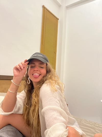 A young woman poses indoors wearing a gray "Balenciaga" baseball cap and a loose, white linen shirt. She sticks her tongue out playfully. Light brown, wavy hair cascades over her shoulder. She wears a silver bracelet on her wrist, along with a white beaded bracelet. A slatted blind hangs on the white wall behind her. The image is rotated, appearing slightly tilted.