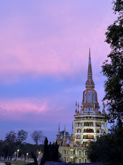 In the city of Ban Khao Tang, Thailand, a towering, multi-tiered pagoda is illuminated against a twilight sky of pastel pink and blue. The pagoda's ornate architecture features gold accents and a central spire. In the foreground and to the right, dark green foliage partially obscures the structure. The surrounding landscape shows other smaller pagodas and trees, with some lights visible, suggesting an outdoor temple complex. The time appears to be just after sunset.