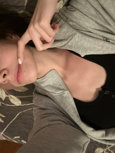 An upside-down selfie featuring a woman lying on a bed. She wears a gray hoodie partially zipped, revealing a black top. Her face is visible from the nose down, and her left hand is near her mouth, with a finger touching her lip. The bed has a patterned gray and white cover.