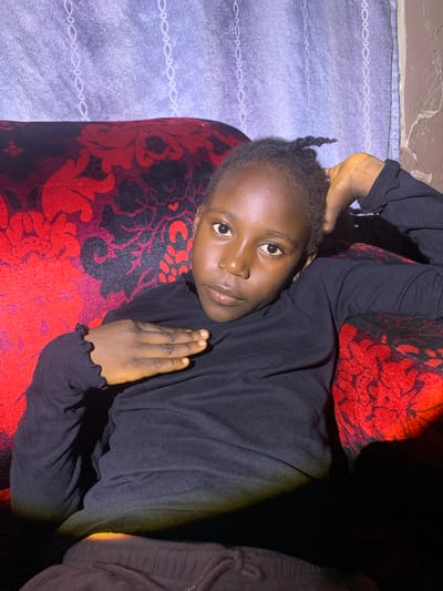 A young Black girl is seated on a red and black patterned sofa, leaning back and resting her head on her right hand, which is propped behind her head. She is wearing a black long-sleeved shirt and dark pants. Her left hand is placed across her chest. The background features a light-colored curtain with a subtle white pattern and a rough, textured wall on the right. The lighting is dim, with a yellow-orange light source casting a glow on the lower portion of the image and on the girl's legs. The overall scene suggests an indoor setting, likely a home, within the city of Barakin Sabawa, Nigeria.