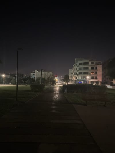 The image presents a nighttime urban scene, likely in Engomi, Lefkoşa, Cyprus, characterized by wet ground suggesting recent rain. A paved pathway, reflecting ambient light, stretches from the foreground into the distance, creating a sense of depth.

To the right of this path, a prominent multi-story building with a distinct curved facade stands, its numerous windows softly illuminated from within. Further down the path, other residential buildings are visible on the right, also displaying interior lights. On the immediate left of the pathway, a darker, possibly grassy area contains a tall, slender utility pole. The path itself is bordered by low-lying bushes or hedges.

In the far background, blurred lights from streetlights and what appear to be distant vehicle lights cast a faint glow against the dark night sky, hinting at an active road or intersection. The overall atmosphere is quiet and still, typical of an urban environment after dark. There are no people visible, nor any discernible text in the frame.