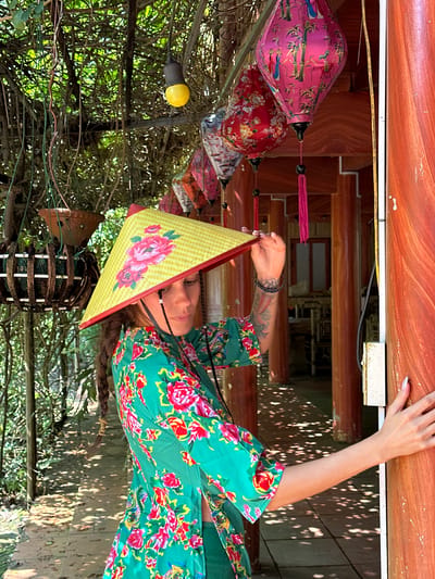 A medium shot captures a woman in an outdoor or semi-outdoor setting, likely in Thông Nguyên Commune, Vietnam. The image is oriented vertically (rotated 90 degrees clockwise from original upload, appearing to be a landscape shot).

The central subject is a woman with long dark hair, wearing a vibrant green garment adorned with large red, pink, and blue floral patterns. She holds a yellow conical hat (Nón Lá) decorated with pink floral motifs, looking towards the camera with a neutral expression. Tattoos are visible on her left forearm. Her right hand rests on a dark wooden beam or railing, which runs across the top of the frame.

The background features multiple colorful Vietnamese silk lanterns hanging to the left of the woman, including a prominent pink one with bamboo and bird patterns, and a red one with floral designs. Above her, a wooden ceiling or roof structure is visible. To her right and behind, dense green foliage creates a natural canopy, with dappled sunlight filtering through the leaves, indicating a sunny daytime scene. Amidst the foliage, a yellow light bulb and a decorative woven wooden object are discernible. The overall setting suggests a charming, culturally rich environment. No visible text is present.