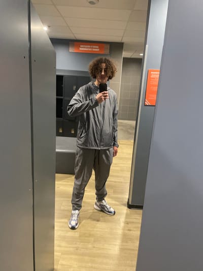 A young individual with curly dark hair, dressed in a gray hooded tracksuit and white, blue, and silver athletic sneakers, is depicted in a mirror selfie. The person stands facing the mirror, holding a black smartphone with the screen visible, and exhibits a slight smile. The scene is a locker room or changing area located in Hérouville-Saint-Clair, France. In the background, gray metal lockers are visible behind a gray bench. The floor is light wood-effect laminate, and the walls are gray. Overhead lighting fixtures are present, and a rectangular orange sign displaying inverted French text is reflected in the mirror.