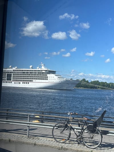 The image depicts a large white cruise ship, identified as "EUROPA 2," docked on a body of water. The ship is positioned horizontally across the frame, with its bow to the left. The water is calm and reflects the blue sky and white clouds. In the foreground, a metal railing lines a waterfront promenade. Two bicycles are parked along the promenade, one with a child seat on the back. Beyond the ship, a line of green trees is visible along the distant shore, suggesting an urban or park setting. The sky is bright blue with scattered cumulus clouds. The overall scene suggests a sunny day in a port city. The specific location is likely a harbor or waterfront area in Stockholm, Sweden, given the context provided.