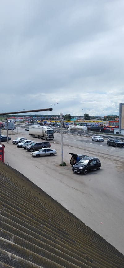 This image depicts a daytime scene in Bulgakovo, Russia, with a focus on a parking area adjacent to a highway. The sky is overcast with a light, grayish-blue hue.
In the foreground, a corrugated, weathered roof covers the lower left portion of the image. A gravel and asphalt parking area is visible, with several cars parked. A dark-colored SUV, possibly a Chery Tiggo, has its trunk open and two individuals are interacting with it. Next to this SUV is a lighter-colored sedan. Further back, a line of parked cars includes a silver sedan, a gray SUV, a white sedan, and a darker sedan.
The highway runs horizontally across the mid-ground of the image. Traffic is visible on the highway, including a large white semi-truck with a trailer. Other vehicles, including sedans and SUVs, are also on the highway.
In the background, a commercial area with various buildings and businesses is visible. Signs with Russian lettering are present on the buildings. The overall impression is one of a busy roadside commercial zone with a highway passing through.
