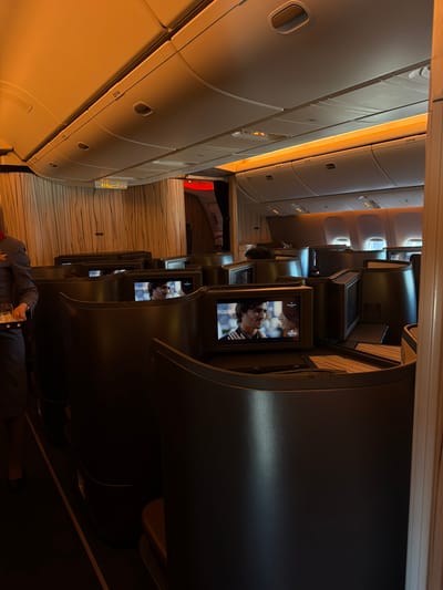 The image shows the interior of an airplane cabin, specifically business or first-class seating. Several rows of seats are visible, each equipped with a personal entertainment screen displaying various images. The overhead compartments are lined with white panels and emergency signs are visible. The general ambiance appears to be warm and softly lit. A flight attendant in uniform and holding a glass stands in the aisle. Location details, such as Taoyuan City, Taiwan, are not discernible within the image itself and cannot be confirmed.