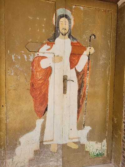 This image depicts a weathered golden-brown double wooden door, likely an exterior entrance, located in Ebenebe, Nigeria. The door features a full-body, hand-painted depiction of Jesus Christ as the Good Shepherd. Jesus is centrally positioned, with dark hair, a beard, and a faint halo. He wears a long white robe and a red cloak draped over his shoulders. His left hand rests on his chest, and his right hand holds a dark, curved shepherd's crook. His bare feet are visible, positioned on painted steps. A white lamb is partially visible to the left of Jesus, also on the painted steps. Green grass is painted at the lower right base of the door. A metallic, tarnished door handle and latch mechanism are centrally located, bisecting the painted figure of Jesus. The door surface exhibits significant paint loss, chipping, and discoloration, particularly on the left panel and around the door handle. A red-brown brick wall is visible on the far right edge of the frame.