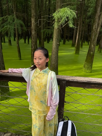 A young East Asian girl with dark hair pulled back in a ponytail, wearing a yellow patterned traditional Chinese dress and a pink and white cardigan, stands on a wooden boardwalk. She is looking to her left, with her left arm resting on the wooden railing. Behind her, a dense forest of tall trees with bare trunks rises from a vibrant green, algae-covered body of water. The water's surface reflects the green of the algae and the dark tones of the trees. A black and white backpack is placed on the boardwalk to the girl's right. The scene appears to be in a natural wetland area within the Lin'an District of China.
