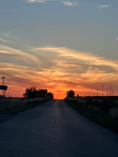 The image depicts a paved road stretching towards a vibrant sunset over a rural landscape. The sky is filled with streaks of orange, yellow, and blue, with wispy clouds. Silhouetted trees and power lines are visible on either side of the road, along with tall grass and dry vegetation. The road itself is dark and shows signs of wear. The scene appears to be located outside the city of Chirpan, Bulgaria, given the characteristic rural setting and the presence of power infrastructure often found in such areas. There are no people or animals visible in the frame.