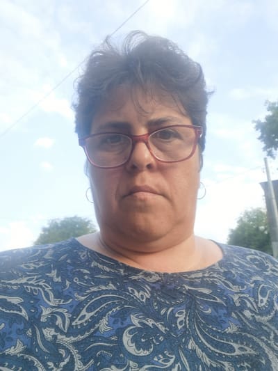 A woman wearing glasses and a blue paisley top stands outdoors, looking directly at the camera. The background shows a sky with clouds, trees, and a utility pole, suggesting an outdoor setting within the city of Lomets, Bulgaria. The lighting indicates it is daytime.