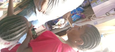 The image, taken in Gayawa, Nigeria, captures a sideways view of an indoor or semi-outdoor setting, likely a small shop or market stall. The scene features three individuals and various packaged goods.

In the foreground on the left, a child's head is visible, adorned with dark cornrow braids. The child wears a light-colored top with a floral pattern. To the right, an adult female is seen in profile, looking towards the left. She also has cornrow braids and is dressed in a vibrant red garment, possibly with green trim. A small earring is visible on her right ear.

Towards the center, partially obscured by the woman, another individual (likely male) is visible from the waist down, wearing blue jeans and a light-colored t-shirt. This person holds a small, clear plastic bottle with a white cap in one hand and a bunch of keys in the other.

The setting includes several commercial products. A large white bag prominently features the text "DANGOTE SUGAR". Nearby, blue packages with "KISSKISS" branding are visible. On a white surface or shelf to the right, small boxes are stacked, bearing the label "Dudu-Osun". White and red electrical cables are also visible on a surface in the background, alongside some wooden structures that may be shelving.

The activity appears to be an everyday interaction or transaction taking place within the shop. The bright lighting suggests it is daytime. The presence of specific branded products like Dangote Sugar, Kisskiss, and Dudu-Osun serves as a strong location cue, consistent with the Nigerian context. Emotions are not clearly discernible from the partial views of the individuals.