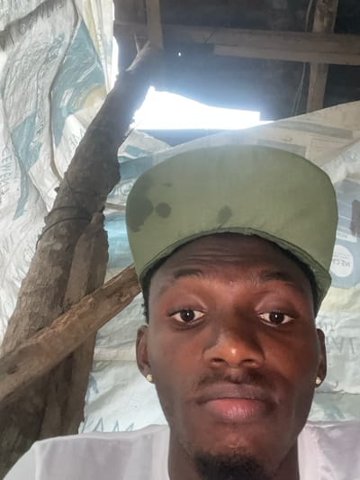 The image is a close-up, selfie-style portrait of a young Black male wearing a green baseball cap and a white t-shirt. He has brown eyes, a mustache, and a goatee. He is wearing a small gold earring in his left ear. The background appears to be an improvised shelter constructed with plastic sheeting and wooden beams. A rough, unworked tree branch is visible to the left of the man, leaning diagonally across the frame. The lighting suggests it is daytime, with a bright light source coming from above and behind the subject, creating some glare and shadows. The overall setting is rustic and appears to be an outdoor or partially enclosed space. The location is specified as Emure-Ekiti, Nigeria.